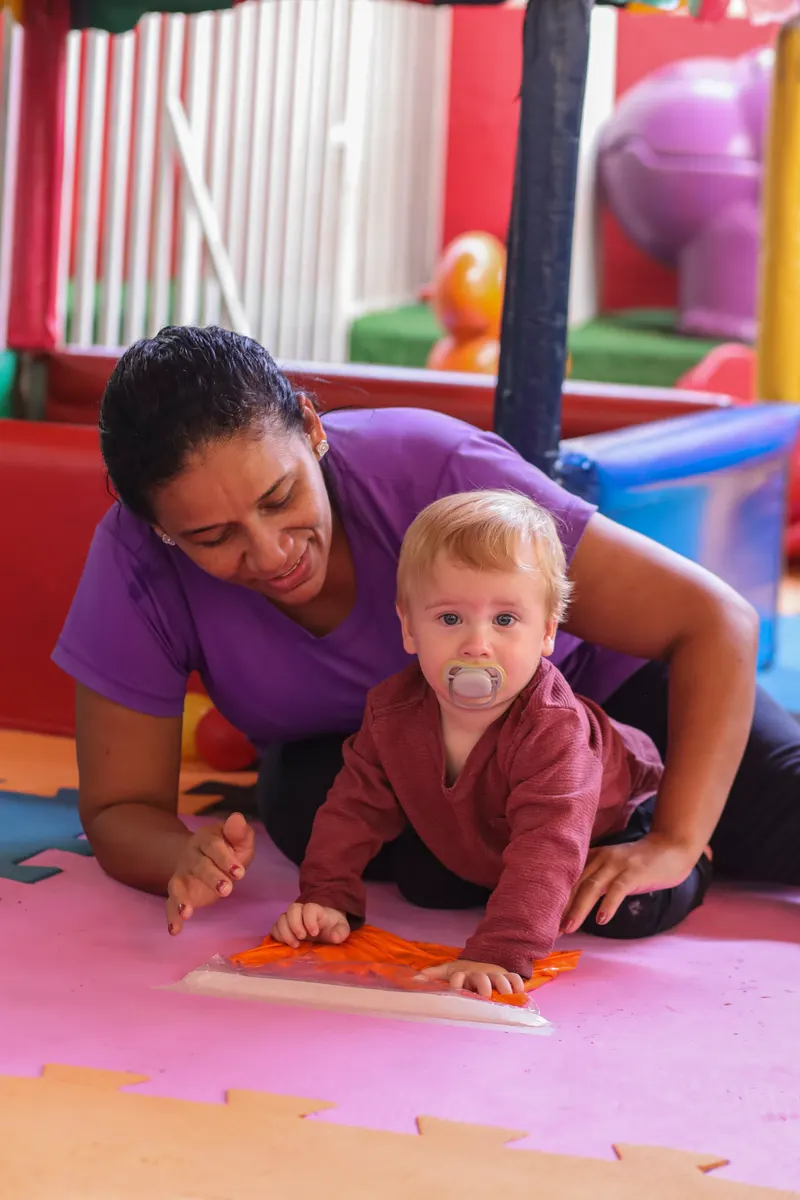 Cuidadora agachada junto a um bebê em atividade sensorial no tapete