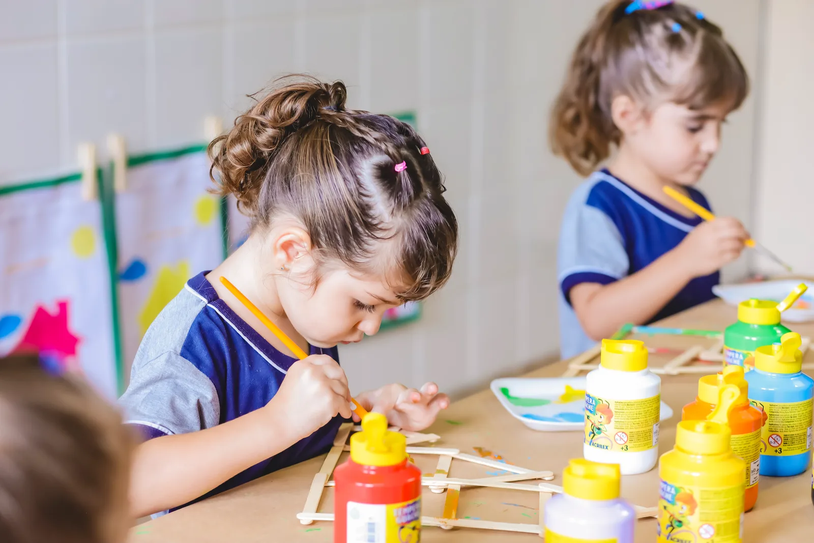 Menina de uniforme Criarte concentrada pintando palitos de sorvete com pincel