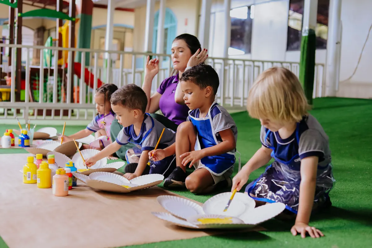 Crianças pintando pratos de papel no chão com a professora