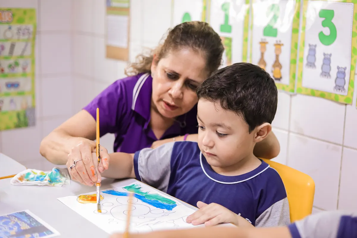 Professora ajudando criança a pintar com tinta guache na mesa