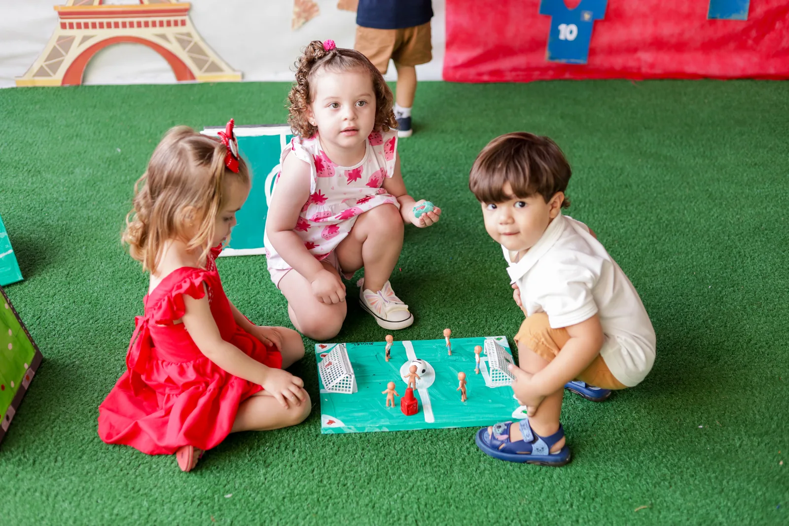 Três crianças da Criarte II na grama, brincando com a maquete de um campo de futebol