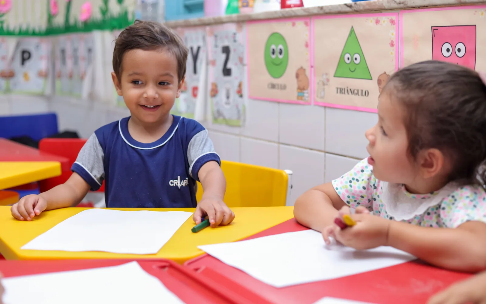 Duas crianças de uniforme Criarte na mesa da sala, com giz de cera e papel, durante a rotina do dia