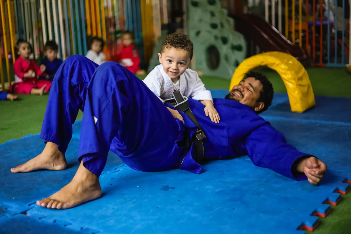 Bebê de quimono branco brincando no tatame com o professor de judô da Criarte